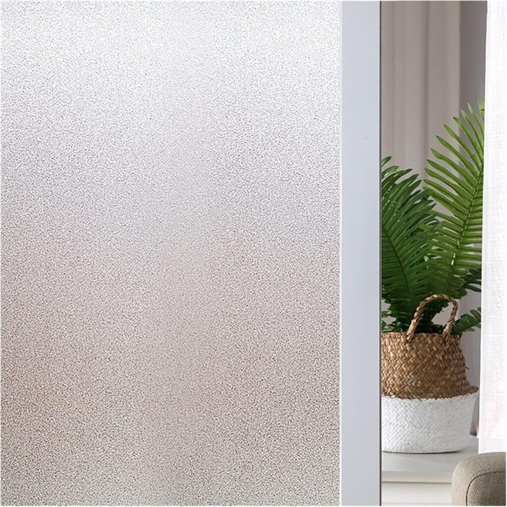 Một tấm kính mờ cát được dán decal che phủ một phần căn phòng. Bên phải, một giỏ đan bằng tre có cây dương xỉ xanh đặt trên bàn gần rèm cửa trắng và ghế bọc vải màu be.